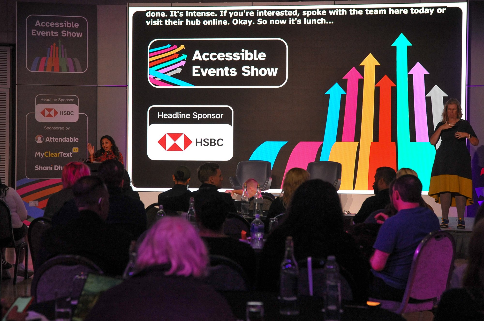 Dr Shani Dhanda on stage at the Accessible Events Show 2025 in front of an audience of event professionals. Live captions and a sign language interpreter are on stage also.